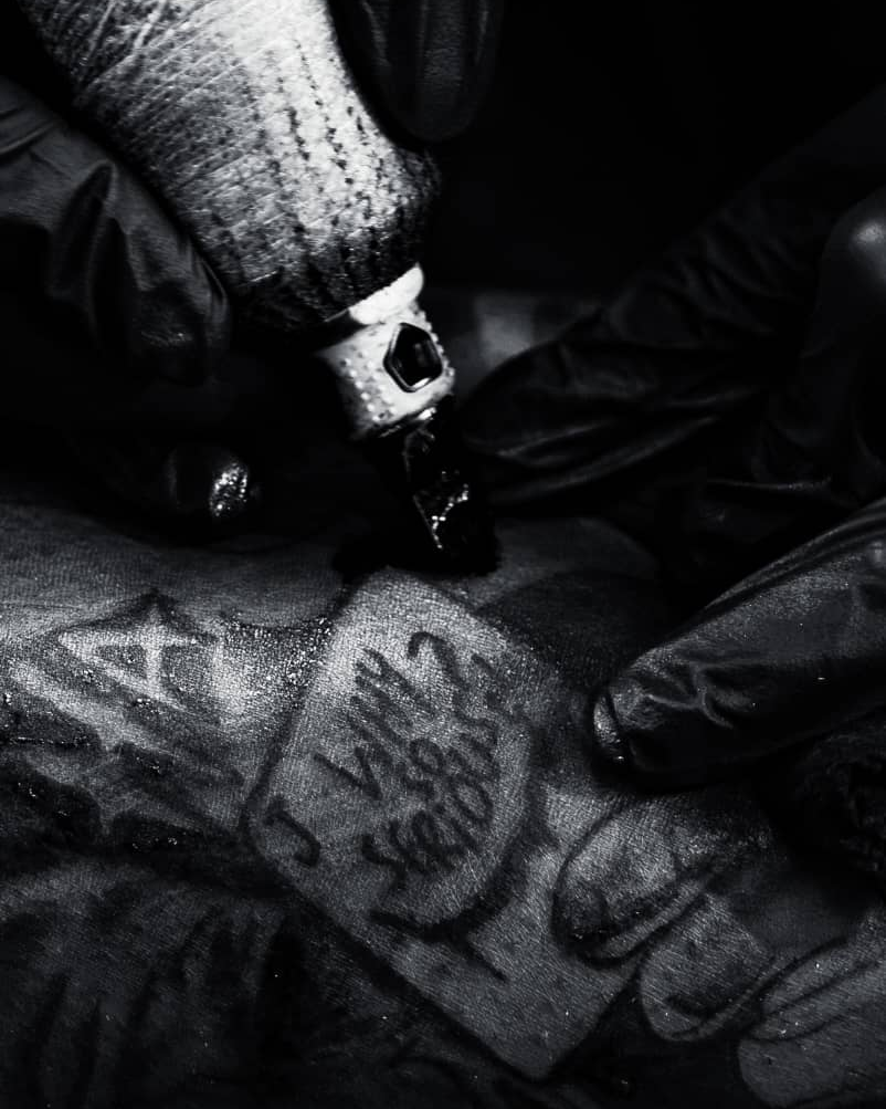 Close-up black and white photo of MAZ tattooing fine line lettering and linework, needle and gloved hands in frame