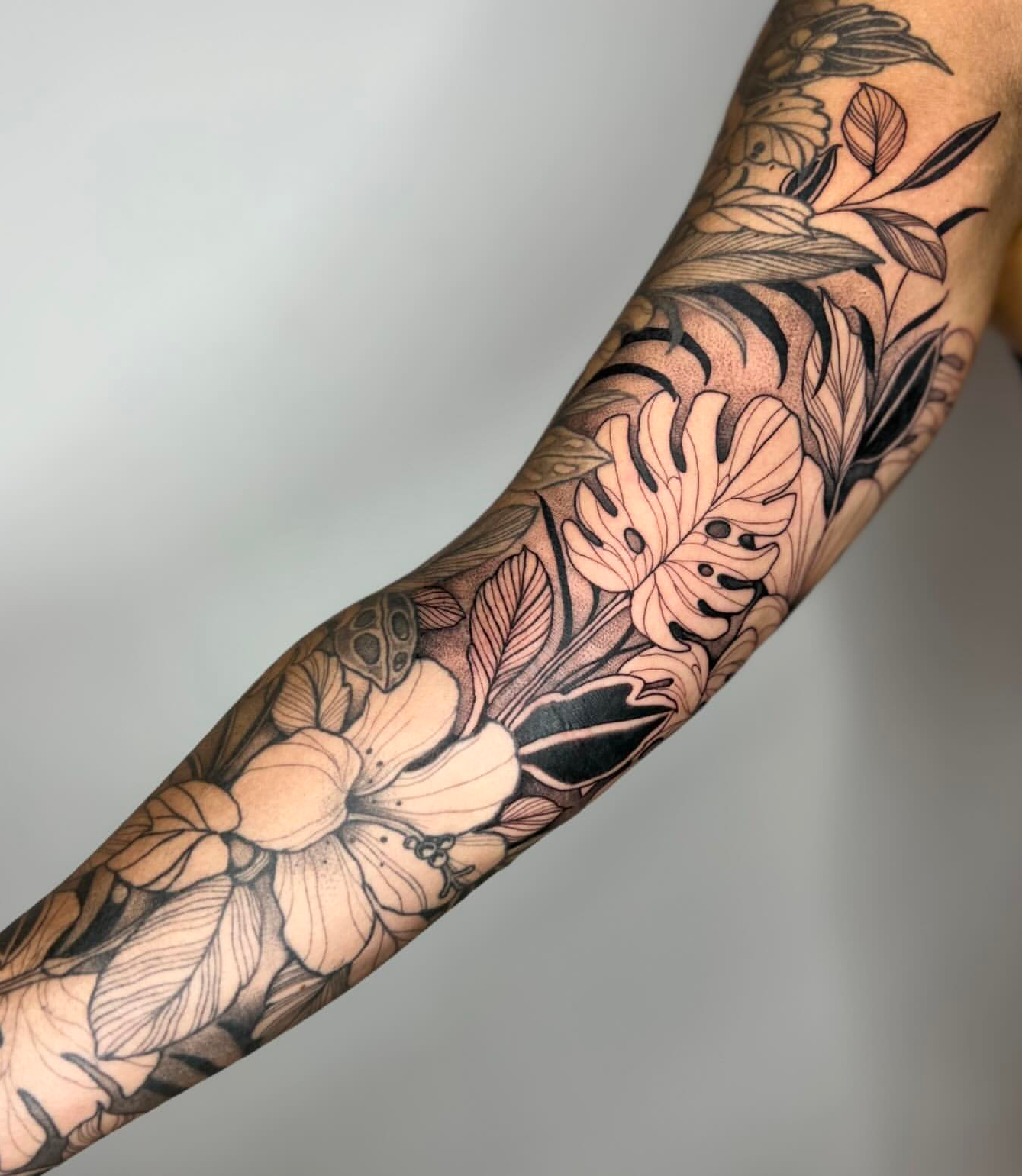 Fine line black and grey tropical botanical forearm sleeve with monstera leaf, hibiscus flowers, and foliage