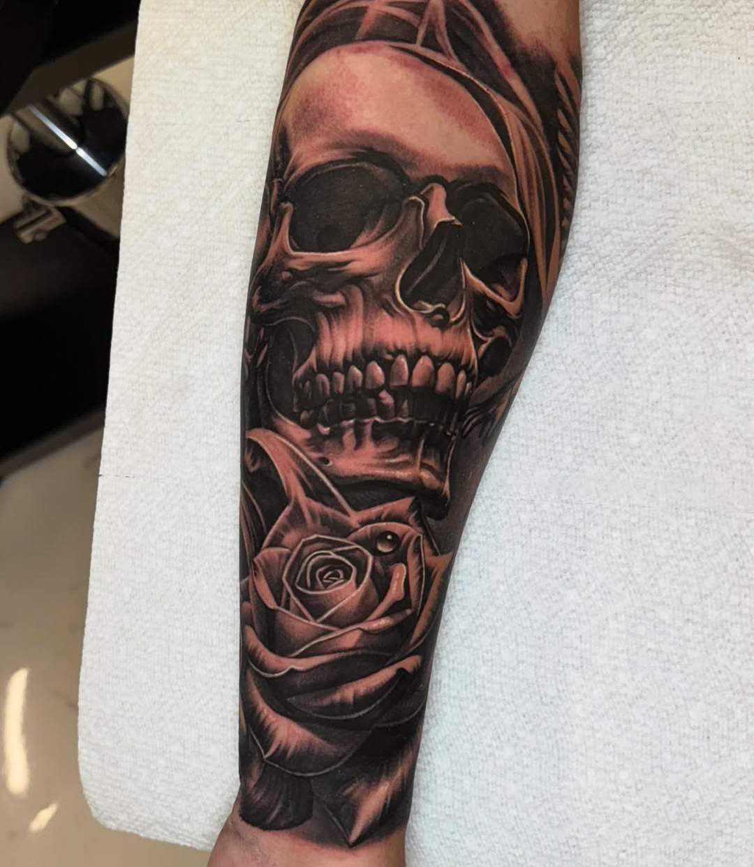 Black and grey realism skull tattoo with hooded shroud and rose, deep gradient shading on forearm