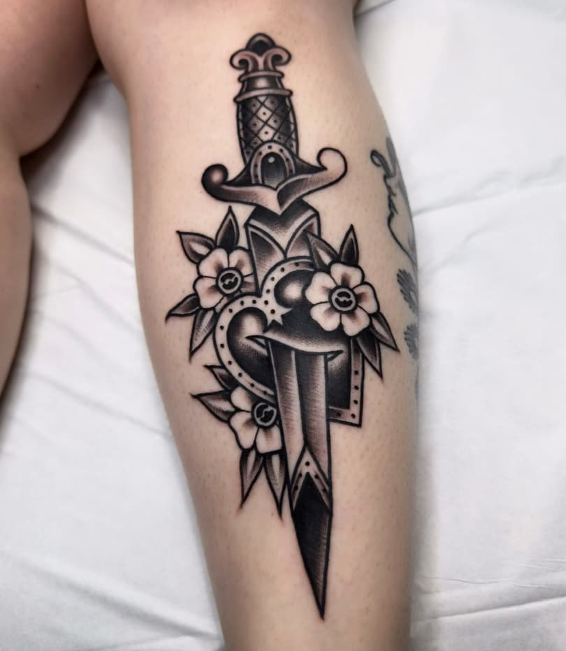 Classic traditional flash dagger tattoo with heart, rope border, and flowers, heavy black outline on calf